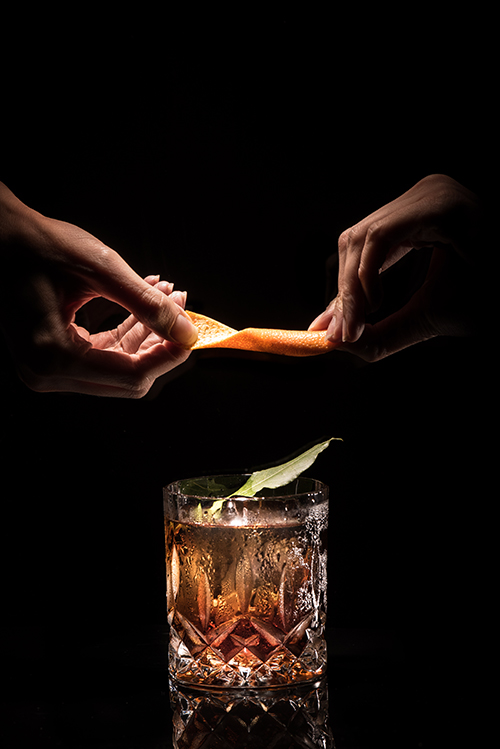 A person squeezing an orange peel into a cocktail glass at The Other Room